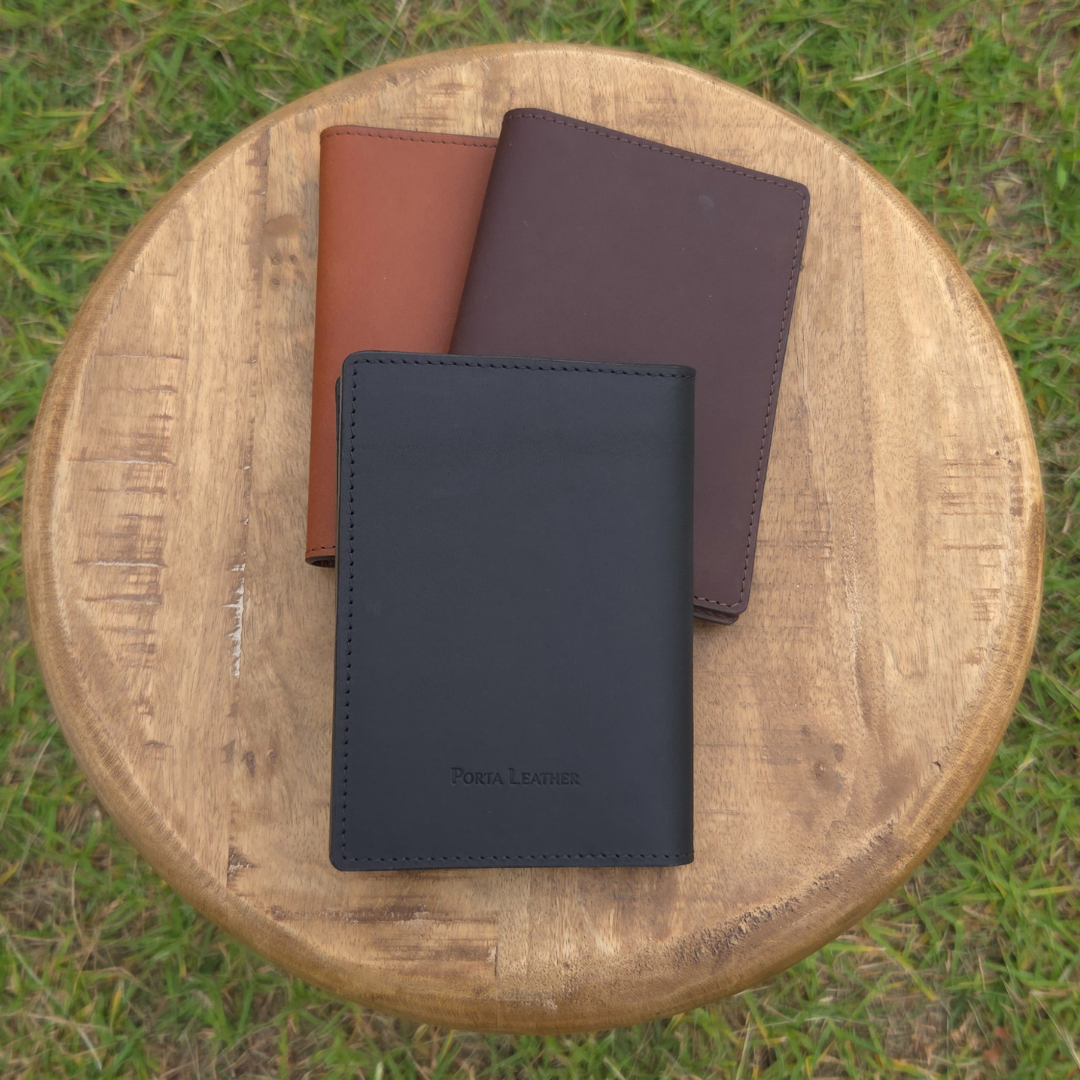 Three Porta Leather wallets in black, dark brown, and brown on a wooden stool outdoors showing the black view of one that has a Porta Leather stamp.