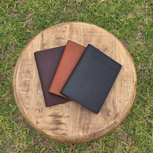 Three Porta Leather passport wallets made from leather in dark brown, black, and brown on a wooden stool outdoors.